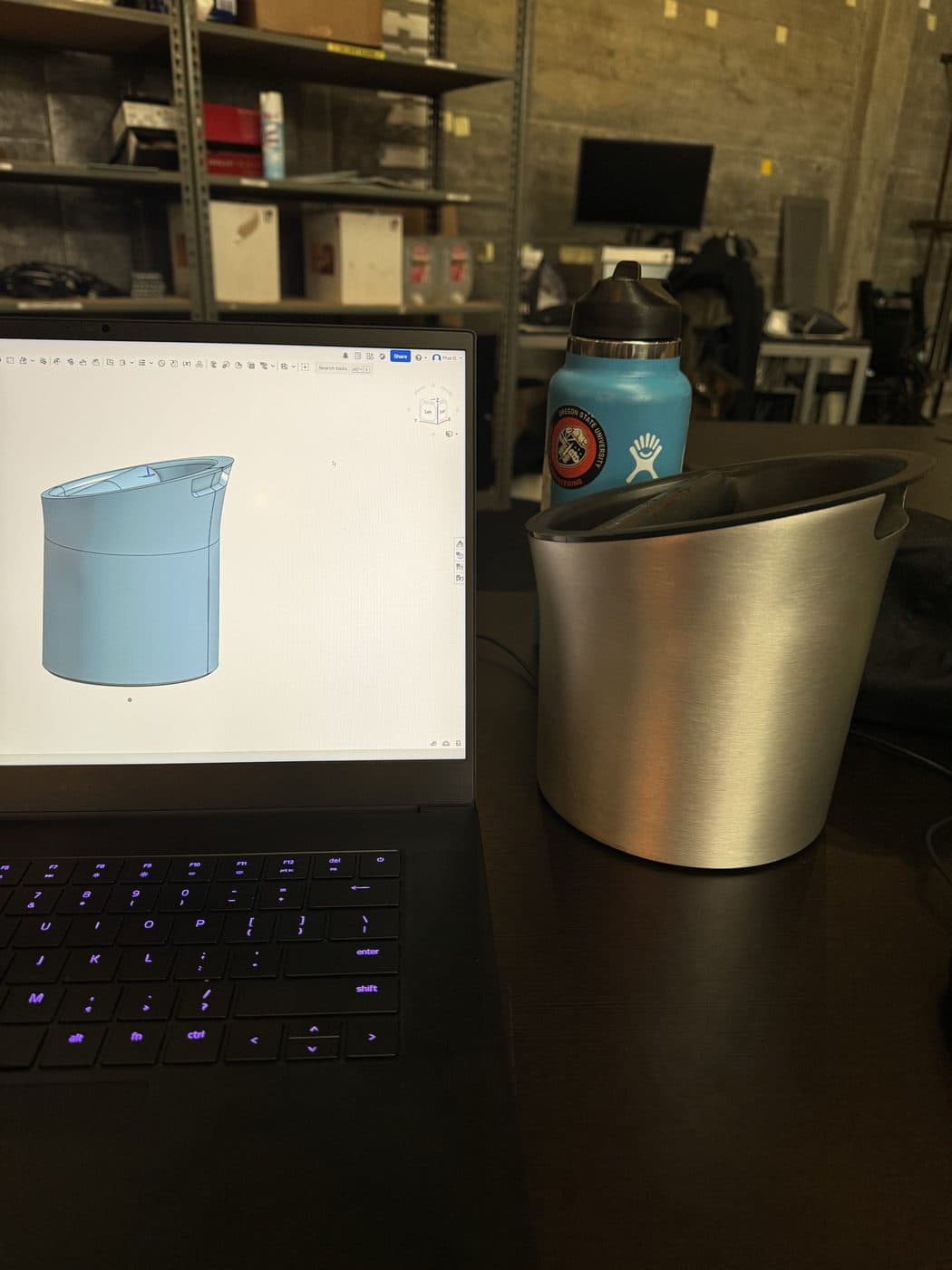 An Onshape model open on the bench next to the formed stainless part it describes.